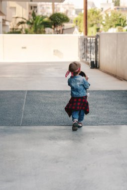 Kot elbise giyen, kırmızı bandana takan küçük çocuk şehir ortamında kaykayla oynuyor..
