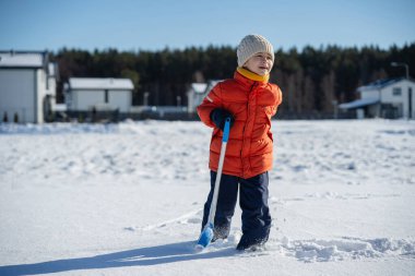 Kışın giyinmiş bir çocuk soğuk havada güneşli bir günde banliyödeki evlerin yakınlarında kar kürüyor..