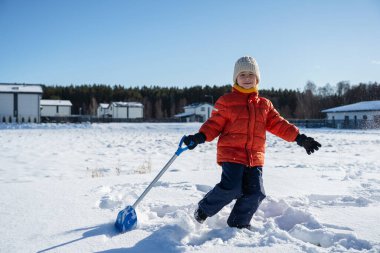 Kışın giyinmiş bir çocuk soğuk havada güneşli bir günde banliyödeki evlerin yakınlarında kar kürüyor..