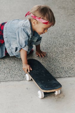 Kot elbise giyen, kırmızı bandana takan küçük çocuk şehir ortamında kaykayla oynuyor..