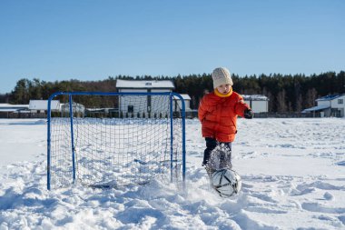 Karla kaplı bahçede kale direğiyle futbol oynayan, kışlık kıyafetler giyen, dışarıda eğlenen ve mevsimlik aktivitelerin tadını çıkaran bir çocuk..