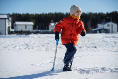 Kışın giyinmiş bir çocuk soğuk havada güneşli bir günde banliyödeki evlerin yakınlarında kar kürüyor..