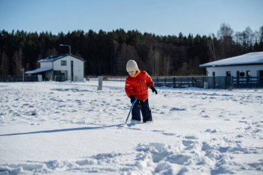Kışın giyinmiş bir çocuk soğuk havada güneşli bir günde banliyödeki evlerin yakınlarında kar kürüyor..