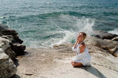 Beyaz elbiseli genç kadın arkasında deniz dalgaları varken kayalık bir kıyıda zarifçe poz veriyor..