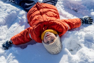 Açık bir kış gününde, açık havada, karlı bir tarlada uzanan çocuk kar meleği yapıyor..