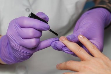 Mor eldivenli profesyonel manikürcü müşterinin tırnaklarına oje sürüyor..
