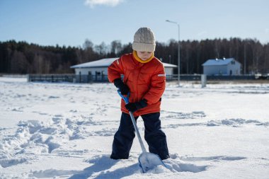 Kışın giyinmiş bir çocuk soğuk havada güneşli bir günde banliyödeki evlerin yakınlarında kar kürüyor..