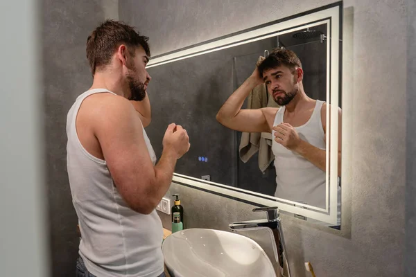 Kolsuz tişörtlü yorgun bir adam banyo aynasında kendini inceliyor..