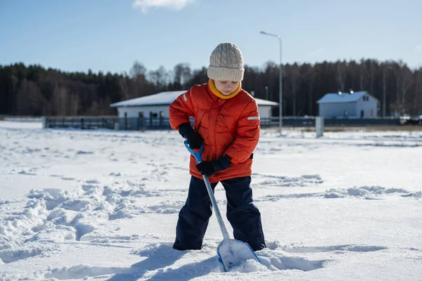 Kışın giyinmiş bir çocuk soğuk havada güneşli bir günde banliyödeki evlerin yakınlarında kar kürüyor..