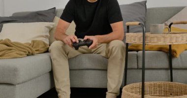 Evdeki kanepede video oyunu oynayan, elinde oyun kumandası olan, gündelik modern oturma odasında dinlenen bir adam..
