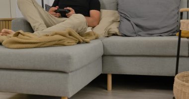Evdeki kanepede video oyunu oynayan, elinde oyun kumandası olan, gündelik modern oturma odasında dinlenen bir adam..