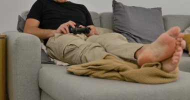 Evdeki kanepede video oyunu oynayan, elinde oyun kumandası olan, gündelik modern oturma odasında dinlenen bir adam..