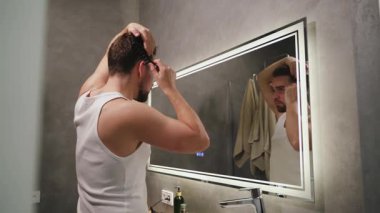 Modern bir banyoda aynaya bakarken saçını tarayan bir adam..