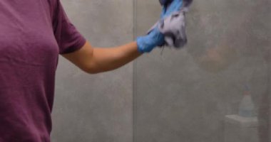 Mavi eldiven giyen bir kadın cam duş duvarını spreyle, kağıt mendille ve cam sileceğiyle temizliyor..