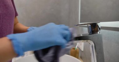 Modern bir banyoda banyo leğeni ve musluğu temizleyen mavi eldivenli kadınların yakın çekim görüntüsü..