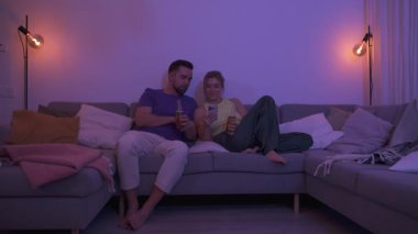 Evdeki rahat bir akşamda içki ve akıllı telefonla koltukta oturan çift..