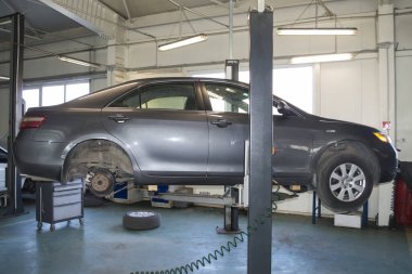 Arka tekerleri sökülmüş gri araba bir oto tamirhanesinde asansöre asılı duruyor.