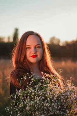 Çekici, kendine güvenen, kızıl saçlı, elinde papatya buketi olan bir kızın portresi. Batan güneş ve parıltı