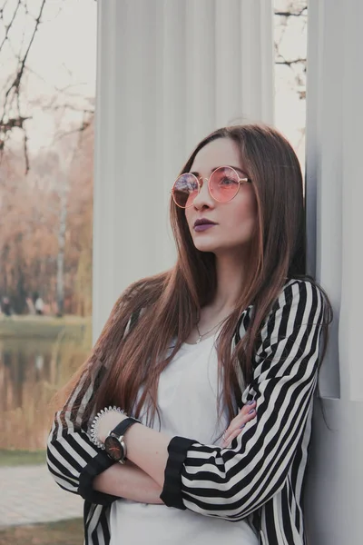 Stylish, confident girl looks into the distance. Pink glasses. Striped black and white jacket. Portrait of a beautiful girl with a strong face. Posing with column