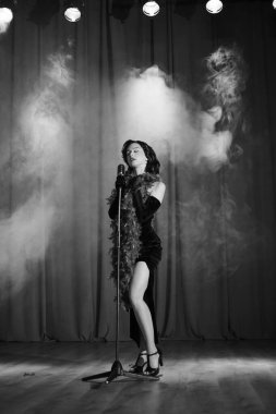 Dansçı sahnede klasik bir mikrofonla dramatik siyah-beyaz bir ortamda performans sergiliyor.