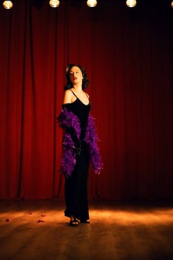 Mor tüylü siyah elbiseli dansçı sahne ışıkları altında bir tiyatro sahnesinde gösteri yapıyor.
