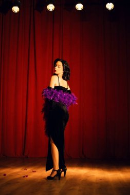 Mor tüylü siyah elbiseli dansçı sahne ışıkları altında bir tiyatro sahnesinde gösteri yapıyor.