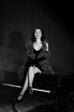 Siyah elbiseli zarif kadın piyanonun üzerinde poz veriyor, klasik tarzını ve loş ışıklandırılmış cazibesini sergiliyor.