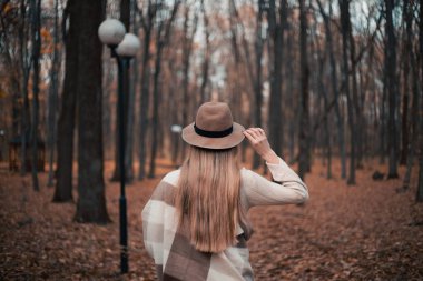 Kahverengi şapkalı bir kadın huzurlu bir sonbahar ormanında yerde yapraklarla yürüyor.
