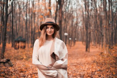 Sıcak kazaklı ve şapkalı bir kadın sonbahar yapraklarıyla dolu bir ormanda gezinti yapıyor.