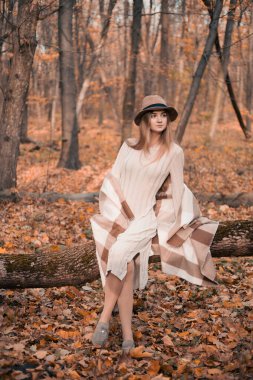 Kadın, sonbaharda bir ormanda düşmüş bir kütüğe zarif bir şekilde oturur. Hafif bir elbise ve şık bir şapka giyer.