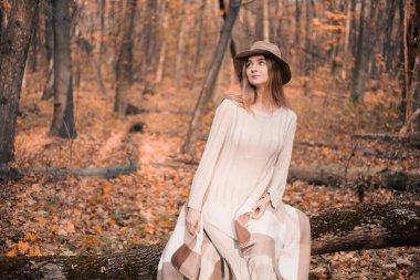 Kadın, sonbaharda bir ormanda düşmüş bir kütüğe zarif bir şekilde oturur. Hafif bir elbise ve şık bir şapka giyer.