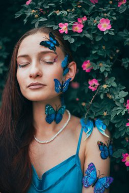 Kelebeklerle dolu yeşil çalıların arasında duran kadın, sıcak bir öğleden sonra sahnesi için bir bahçede poz veriyor.