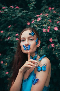 Cildi ve yüzü kelebeklerle kaplı bir kadın gündüzleri çiçeklerin arasında duruyor.