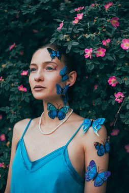 Parlak gün ışığında teninde kelebekler ve arka planında çiçeklerle bahçede poz verir.
