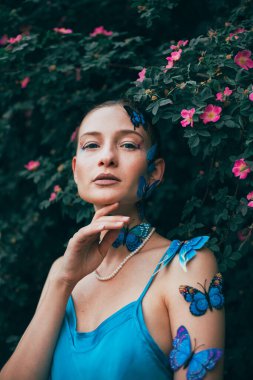 Parlak gün ışığında teninde kelebekler ve arka planında çiçeklerle bahçede poz verir.