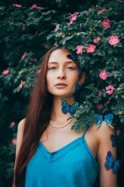 Kelebeklerle dolu yeşil çalıların arasında duran kadın, sıcak bir öğleden sonra sahnesi için bir bahçede poz veriyor.