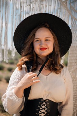 Genç bir kadın açık hava ortamında elinde bir kumaş yayı, üzerinde şapka ve elbiseyle, güneşli havalarda doğal bir manzarayla dikiliyor.