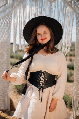 Genç bir kadın açık hava ortamında elinde bir kumaş yayı, üzerinde şapka ve elbiseyle, güneşli havalarda doğal bir manzarayla dikiliyor.