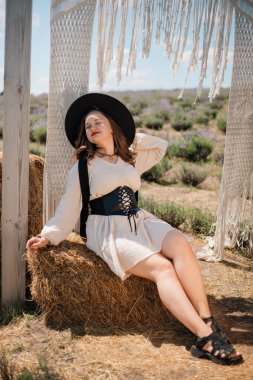 Genç bir kadın açık hava ortamında elinde bir kumaş yayı, üzerinde şapka ve elbiseyle, güneşli havalarda doğal bir manzarayla dikiliyor.