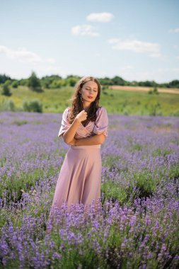 Güneşli bir günde kadın lavanta tarlasında duruyor elleri saçlarında ve nazik bir gülümsemeyle