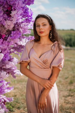 Uzun elbiseli bir kadın güneşli bir günde açık havada mor bir çiçek depolarının yanında poz veriyor.
