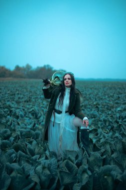 Kadınlar bulutlu bir günde uzun beyaz bir elbise ve yeşil bir palto giyerken tarlada lahana taşırlar.