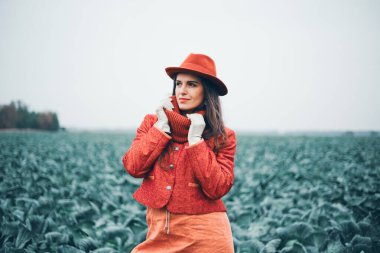 Kırmızı paltolu ve şapkalı kadın bulutlu bir günde yeşil tarlada duruyor.