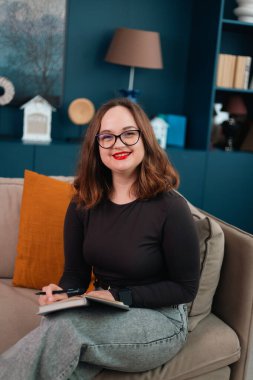 Psikolog kanepede oturur ve mavi raflı ve dekoratif eşyalarla oturma odasında notlar alır.