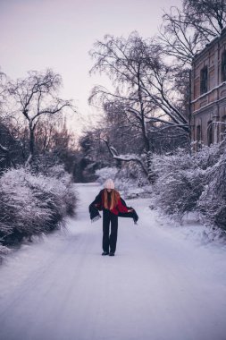 Uzun saçlı ve koyu renk elbiseli bir kadın, kış sahnesinde etrafını saran ağaçlarla karlı bir yolda duruyor.