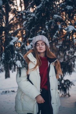 Genç bir kadın kış günbatımında karla kaplı ormanda duruyor. Sıcak kıyafetler giyiyor ve anın tadını çıkarıyor.