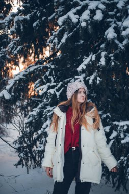 Genç bir kadın kış günbatımında karla kaplı ormanda duruyor. Sıcak kıyafetler giyiyor ve anın tadını çıkarıyor.
