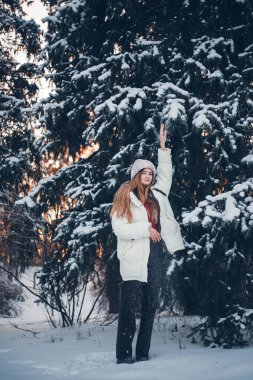 Genç bir kadın kış günbatımında karla kaplı ormanda duruyor. Sıcak kıyafetler giyiyor ve anın tadını çıkarıyor.