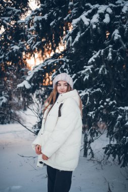 Genç bir kadın kış günbatımında karla kaplı ormanda duruyor. Sıcak kıyafetler giyiyor ve anın tadını çıkarıyor.