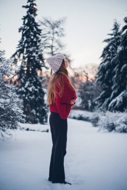 Kadın kışın ağaçlarla karlı bir arazide duruyor, sıcak giysiler ve gün boyunca örülmüş bir şapka giyiyor.